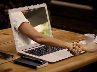 A person working on a laptop shaking hand with an online friend through the laptop screen