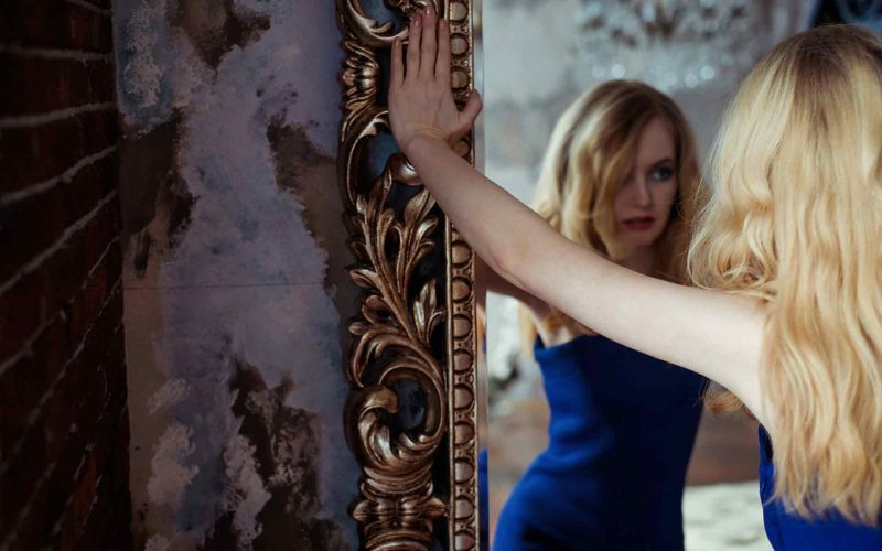A blonde narcissist woman standing and viewing herself in a mirror