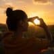 Image of a woman at sunset making a heart sign with her fingers encircling the sun to show self-love.